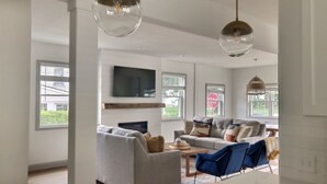 A spacious living room with a grey sofa, blue armchair, and a coffee table. There are large windows providing natural light and a flat-screen TV mounted on the wall. - Rock House - Lake Views & 15 Mins to Cedar Point (Huron)