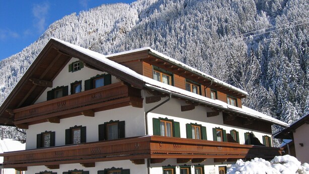 Una cabaña tradicional con techo inclinado, vigas de madera y contraventanas verdes. Está rodeada de árboles nevados y montañas.