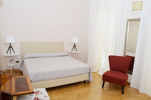 Quarto de hotel com cama, duas luminárias de cabeceira, uma cadeira vermelha, uma mesa pequena e um espelho.
