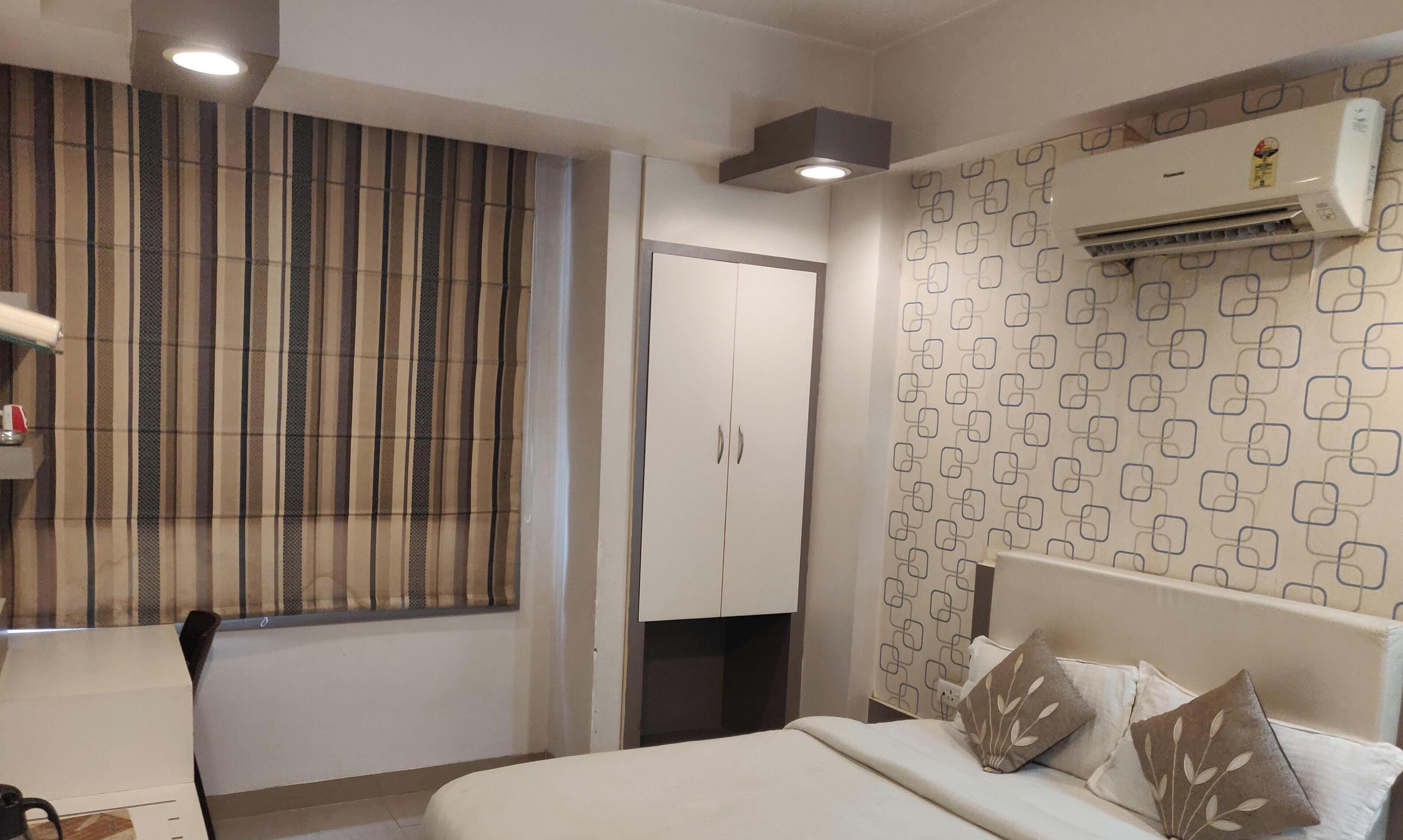 A hotel room with a bed, a wardrobe, a window with curtains, an air conditioning unit, and a patterned wall.