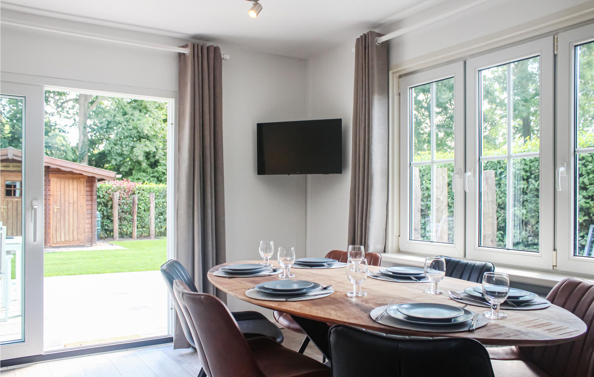 Un comedor con una mesa grande dispuesta para seis personas, un televisor de pantalla plana montado en la pared y ventanas amplias que dan a un jardín.