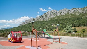 Un parque infantil con un módulo de juego rojo, un tobogán verde y una red de escalada, frente a un paisaje montañoso.