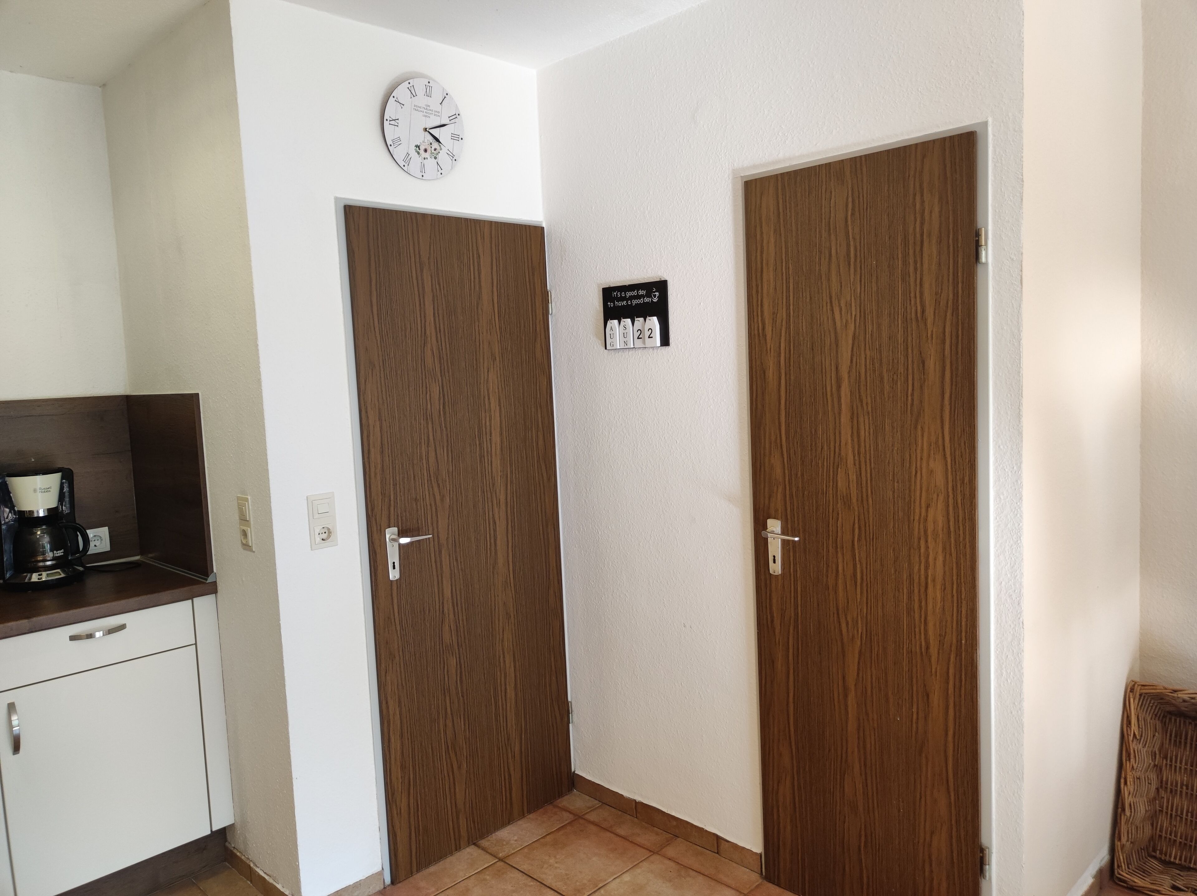 Een hotelkamer met een houten deur, een wandklok, een koffiezetapparaat op een aanrecht, en een bord met een bericht.