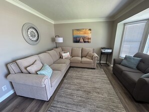 A living room with a beige sectional sofa, a smaller sofa, a rug, a wall clock, a lamp, and a painting.