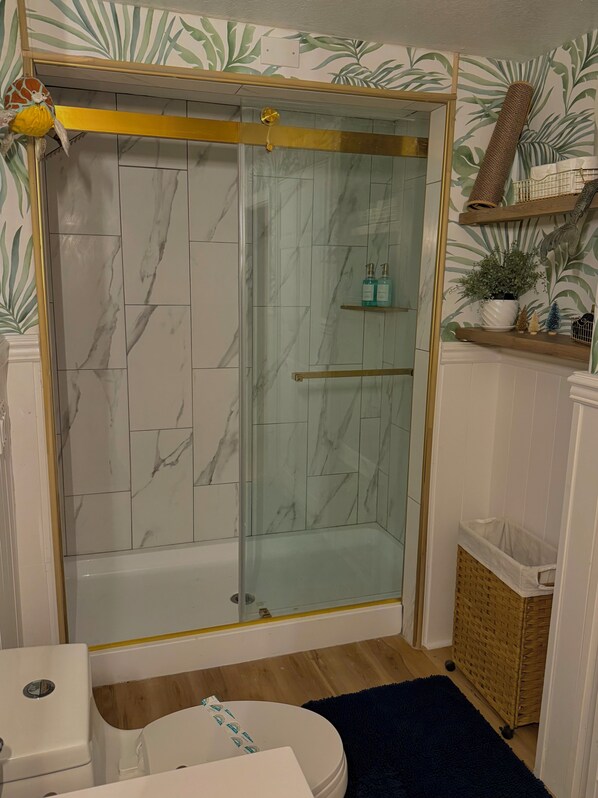 A bathroom with a glass-enclosed shower, marble tiles, a wooden floor, a white toilet, and a blue rug.