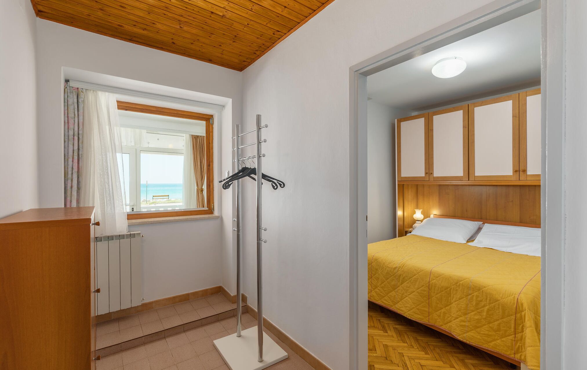 Un dormitorio con cama, armario de madera, radiador y ventana con vista al mar.