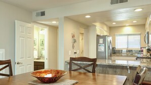 A dining area with a wooden table, chairs, and a large bowl. There is a kitchen in the background with a refrigerator and a window with blinds. - 110 Pismo Shores: 2  BR, 1.5  BA Condominium in Pismo Beach, Sleeps 6 (Pismo Beach)