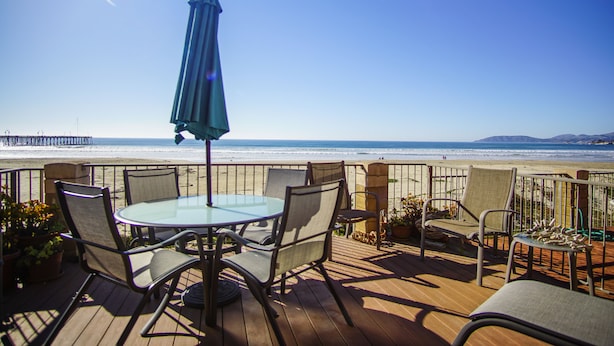 Varanda com mesa e cadeiras de vidro, com vista para uma praia e um píer ao longe.