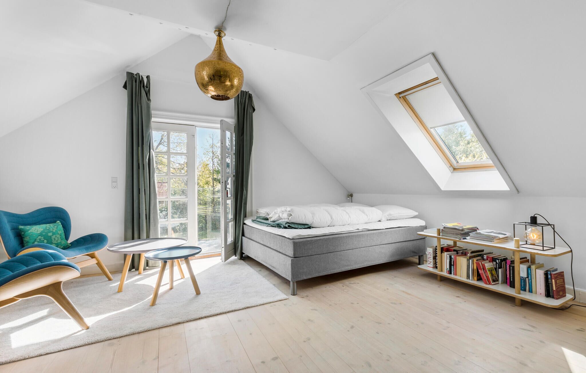 Une chambre de style loft moderne, dotée d’une lucarne, d’un lit, d’une chaise bleue, d’une petite table et d’une bibliothèque.