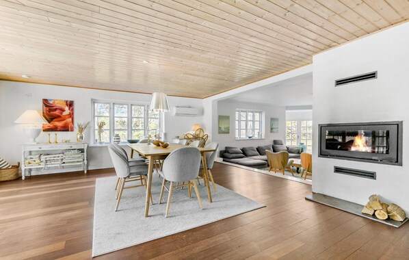 A spacious living area with a wooden ceiling, a dining table with chairs, a sofa, a fireplace, and large windows.