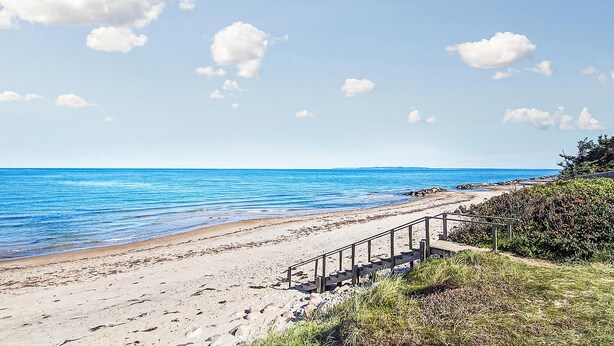 Una spiaggia con una passerella in legno che conduce alla riva, acqua cristallina e una zona verde con cespugli.