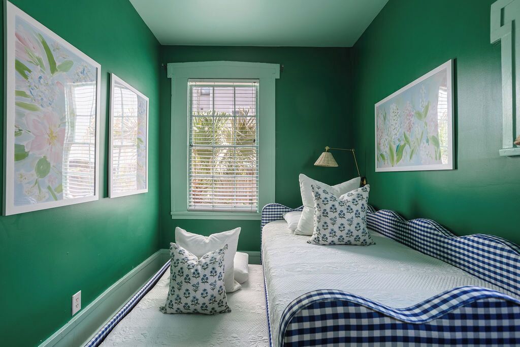 Een kamer met groene wanden, een raam met zonwering, een bed met geruit beddengoed en bloemistisch kunstwerk.