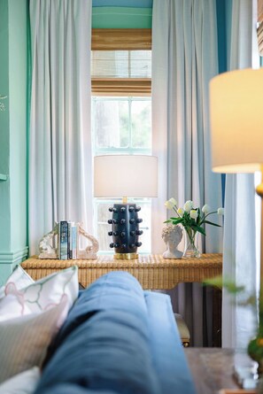 Una habitación con una cama, una mesita de mimbre, una lámpara y una ventana con cortinas.