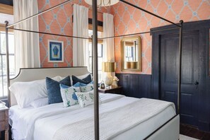 Una cama con dosel, ropa de cama blanca y almohadas decorativas, una mesita de noche con una lámpara y una puerta de madera oscura.