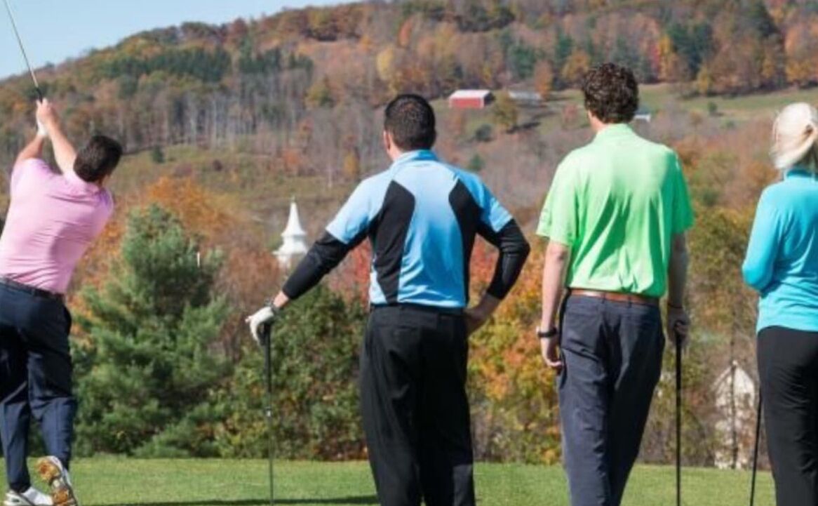 A golf course with players in motion, a clear sky, and autumn foliage in the background.