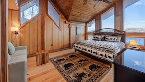 A wooden cabin bedroom with a bed, a chair, a ceiling fan, and large windows. - THE LUXURY LODGE - SKI, RIDE, GOLF, BIKE, HIKE (Hunter)