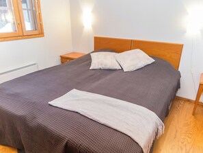 A neatly made bed with a grey bedspread and two pillows, a wooden headboard, a small wooden nightstand with a lamp, and a radiator on the wall. - Fore camp by Interhome (Nilsiä)