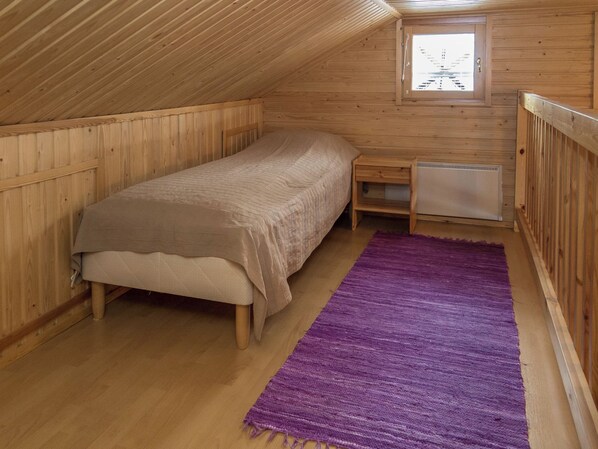 Een eenpersoonsbed met hoofdeinde, een klein nachtkastje, een verwarming en een paars vloerkleed in een kamer met houten wanden en een schuine kap.
