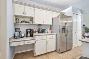 Cocina compacta con armarios blancos, refrigerador de acero inoxidable, cafetera y licuadora sobre la encimera.