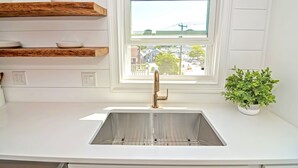 Una cocina con armarios blancos, un lavamanos de acero inoxidable, estantes de madera y una ventana con vistas a edificios y una línea eléctrica.