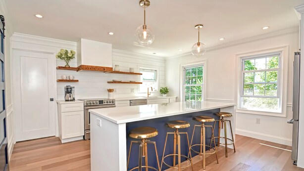 Una cocina moderna con armarios blancos, una isla central con taburetes, iluminación colgante y un amplio ventanal que proporciona luz natural.