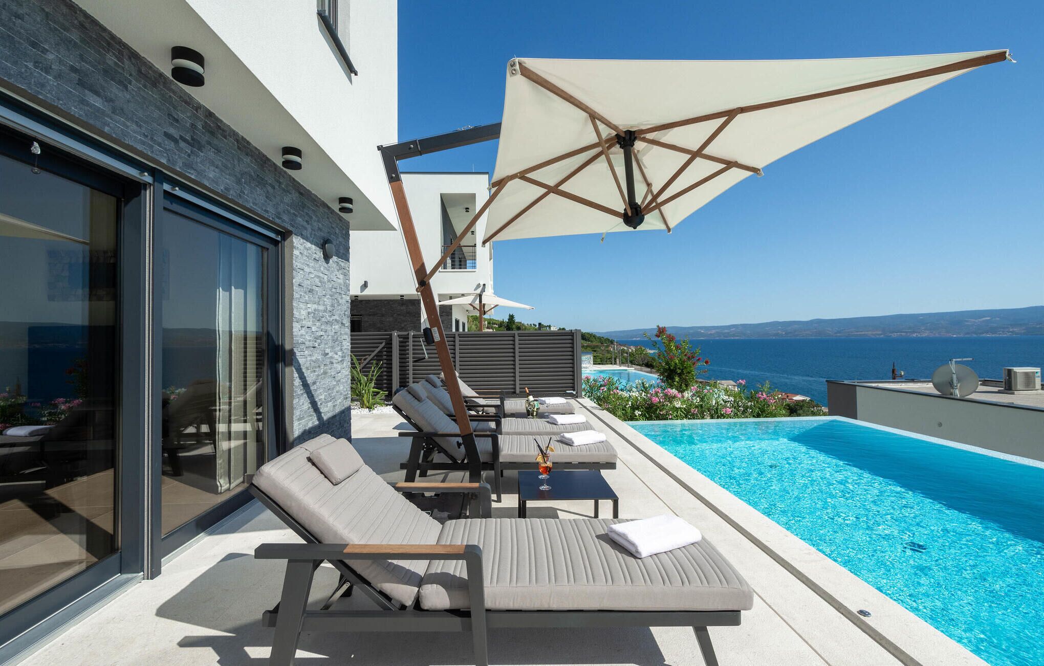 Area piscina moderna all'aperto con lettini e un ombrellone grande, con vista su un mare cristallino e azzurro.