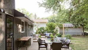 En udendørs terrasse med et glastilbord og fletstole, en træbænk og et lille bord med en vase.