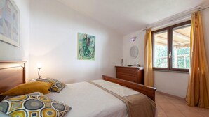 A bedroom with a bed, a dresser, a window with curtains, a painting on the wall, and a nightstand with a lamp.