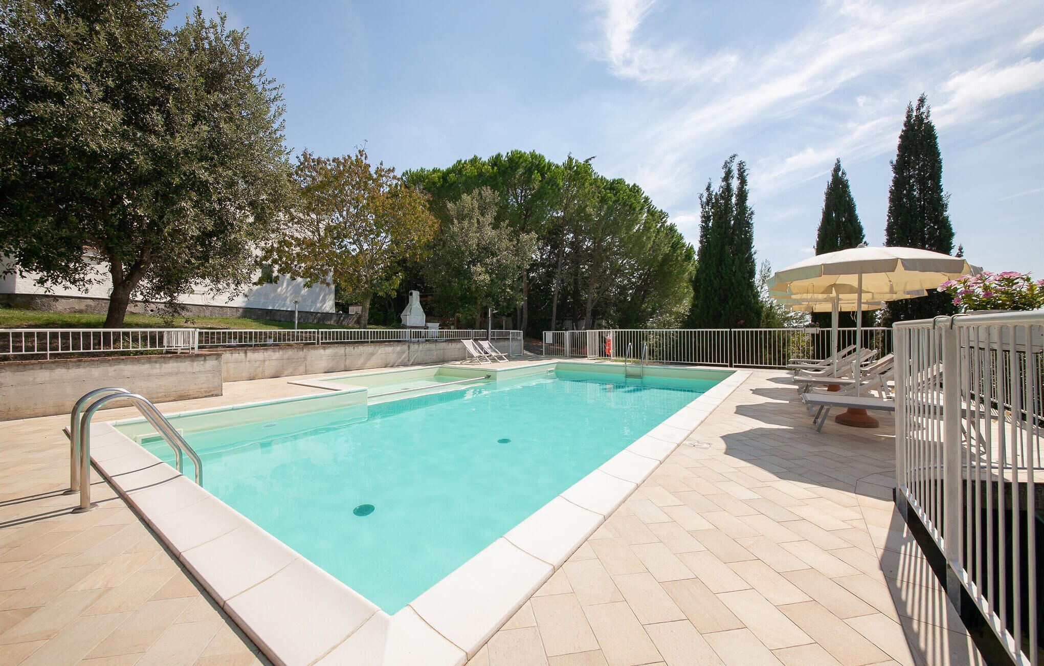 A rectangular swimming pool with a tiled deck, surrounded by a metal fence. There are lounge chairs and a covered seating area with a canopy.