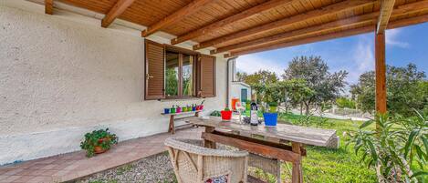Un patio techado al aire libre con una mesa de madera y sillas de mimbre, una ventana con contraventanas y vistas a una piscina y follaje.