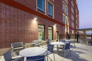 An outdoor patio with round tables and chairs, a brick building with large windows, and a black metal fence. - La Quinta Inn & Suites by Wyndham Dallas - Frisco Stadium (Frisco)