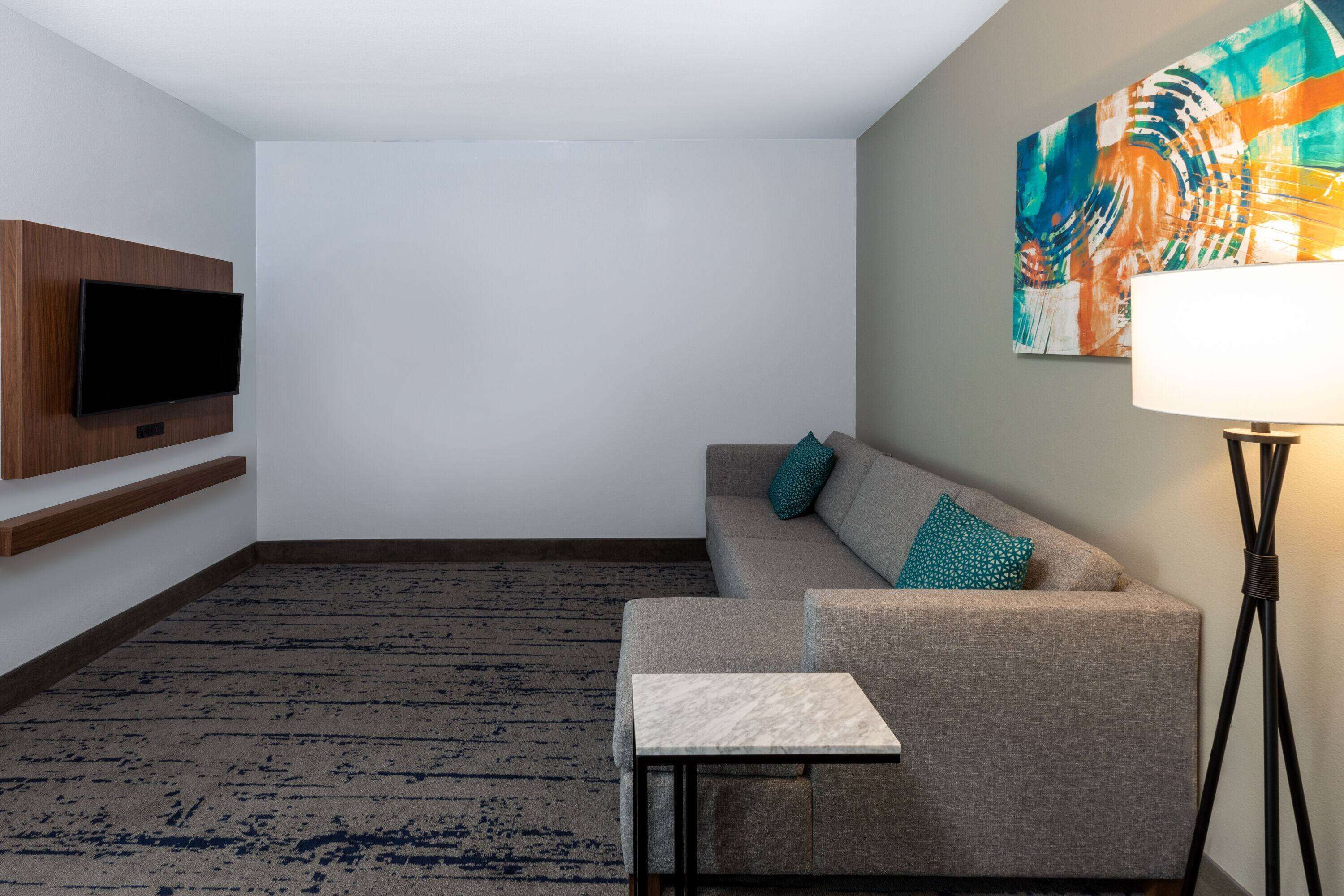 Une chambre d’hôtel moderne avec un canapé, une petite table, un téléviseur à écran plat fixé au mur, une lampe sur pied et une peinture abstraite aux couleurs vives accrochée au mur.