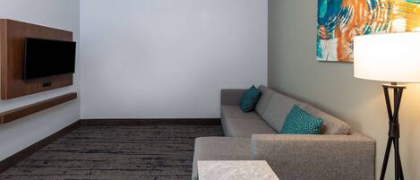 Une chambre d’hôtel moderne avec un canapé, une petite table, un téléviseur à écran plat fixé au mur, une lampe sur pied et une peinture abstraite aux couleurs vives accrochée au mur.