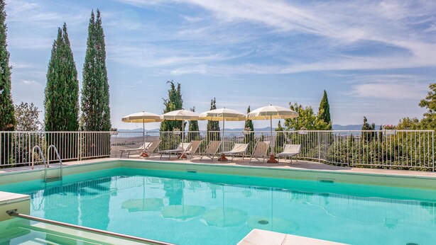 Ein Pool mit klarem, blauem Wasser, umgeben von Liegestühlen und Sonnenschirmen, mit hohen Bäumen im Hintergrund.