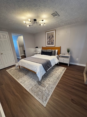 Une chambre à coucher comprenant un lit, des tables de chevet, une lampe, un luminaire au plafond, un tableau encadré et une porte.