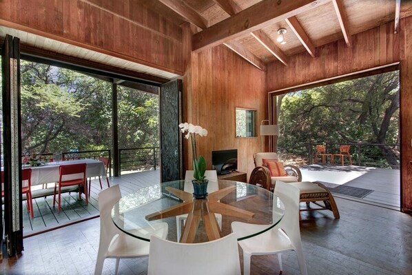 A modern interior with a glass table surrounded by white chairs, a wooden ceiling, and large windows offering a view of trees. - Idyllic Setting on Waialea Bay - White Sand Beach, Family Friendly (Kamuela)