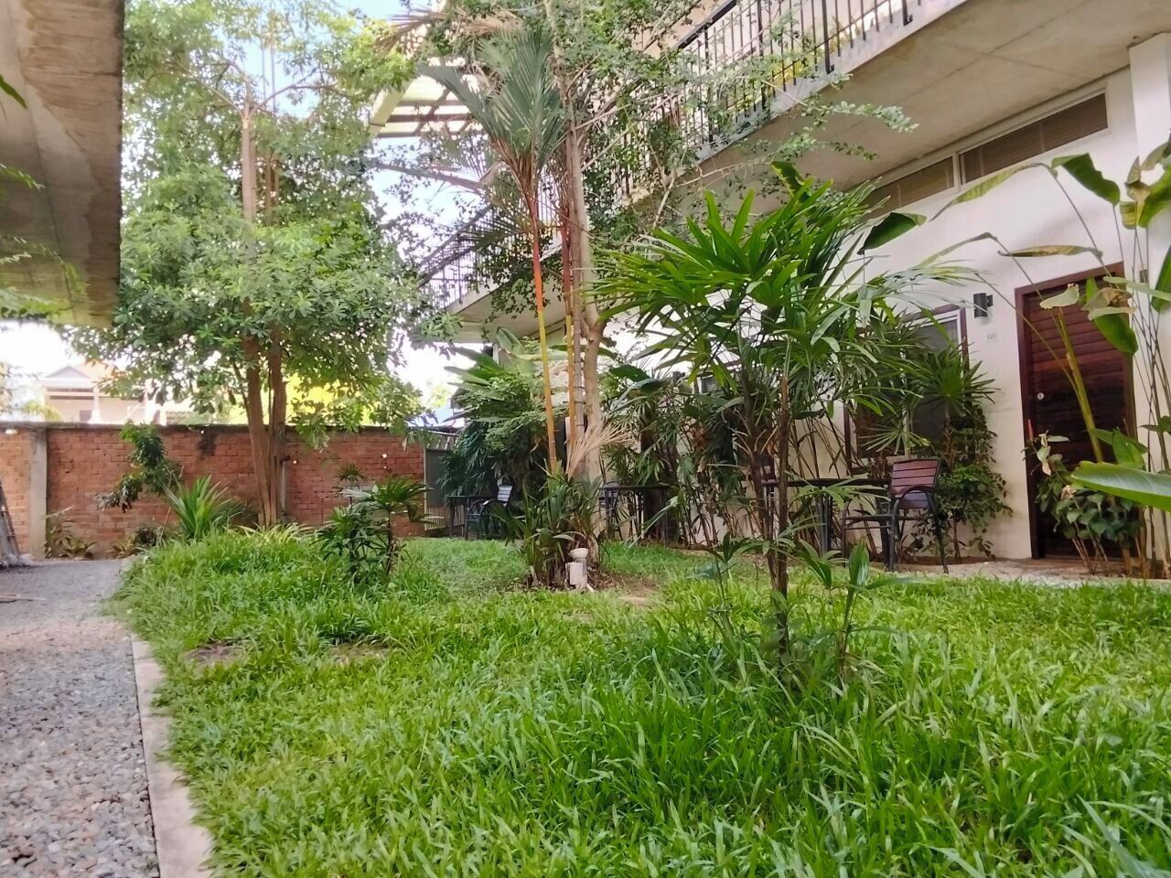 A courtyard with a gravel path, green grass, and various plants. There is a building with a balcony and a door visible.