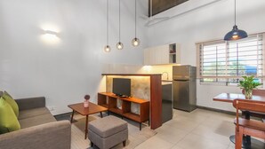 Een moderne woonkamer met een bank, poef en een klein tafeltje. Er is een kitchenette met een koelkast en een televisie.