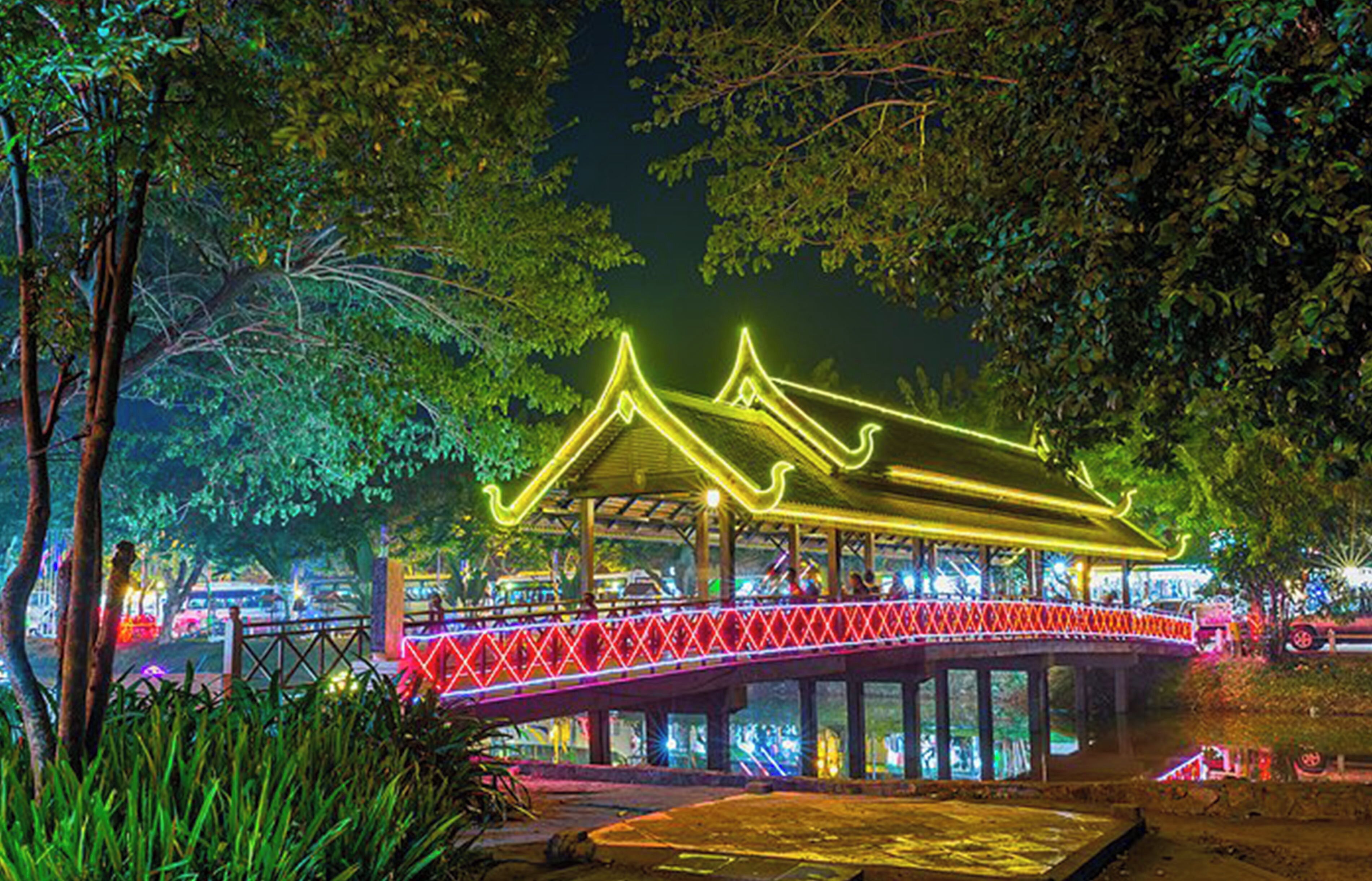 Een klassiek paviljoen met een dak dat rood en geel oplicht met neonverlichting, gelegen op een brug over water, omringd door groen.