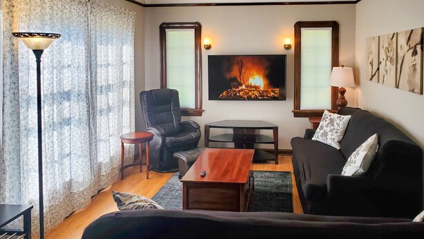 Ein Wohnzimmer mit einer schwarzen Ledersofa, einem Holztisch, einem schwarzen Lederstuhl, einem kleinen runden Tisch und einer Stehlampe. Ein Kamin mit Feuer und zwei Wandleuchten sind ebenfalls vorhanden.