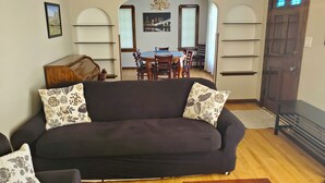 Ein Wohnzimmer mit einer dunklen Couch, gemusterten Kissen, einem Holztisch und einem Essbereich im Hintergrund.