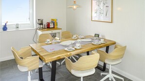 Ruokailutilassa on puinen pöytä, johon on katettu neljälle, ja pöydän ympärillä on valkoisia tuoleja, joissa on pehmustetut istuimet. Pienellä pöydällä ikkunan vieressä on kahvinkeitin, leivänpaahdin ja Novasol-tuotepakkaus.