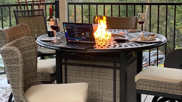 An outdoor patio with wicker furniture, a round table with a laptop and a lit fire, and a view of trees and hills. - Vista Chalet-Yosemite and Bass Lake getaway with Tesla charger (Oakhurst)