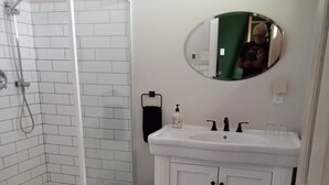 Un baño con ducha de azulejos blancos, un lavabo blanco con espejo y un cubo de basura negro.