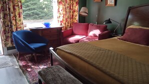 Un dormitorio con una cama, un sofá rojo, un sillón azul, una mesita y una ventana con cortinas.