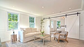 A spacious living room with a large sofa, a coffee table, and a wooden chair. There are large windows allowing natural light and a view of greenery outside. - Harper House Downtown Mystic Romantic Weekend Getaway (Mystic)