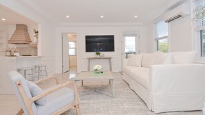 A modern living room with a white sofa, a wooden chair, a coffee table, and a TV mounted on the wall. - Harper House Downtown Mystic Romantic Weekend Getaway (Mystic)