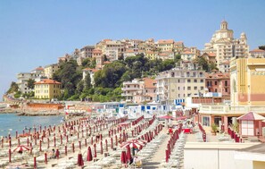 Un hôtel situé sur la côte, avec plusieurs étages, une plage aménagée avec des rangées de transats et de parasols, et une église imposante en arrière-plan.