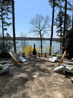 Un patio de piedra con sillas Adirondack y un brasero, con vista a un lago que incluye un bote y árboles al fondo.