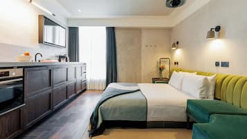 A modern hotel room with a kitchen area, a bed with green upholstered headboard, a bedside table with a vase, and a large window with curtains.